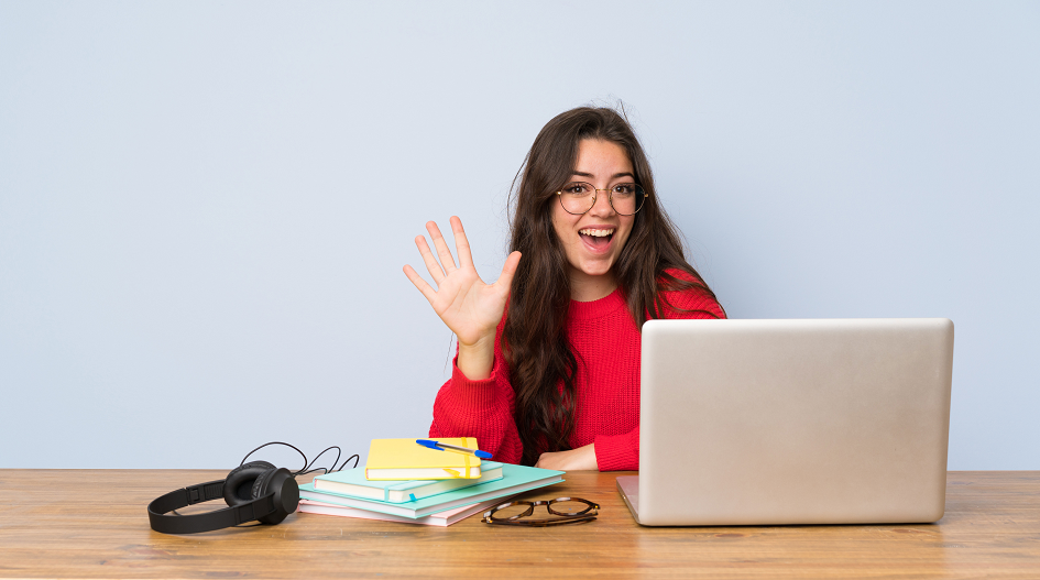 Eine Studentin sitzt an ihrem Laptop und winkt in die Kamera.